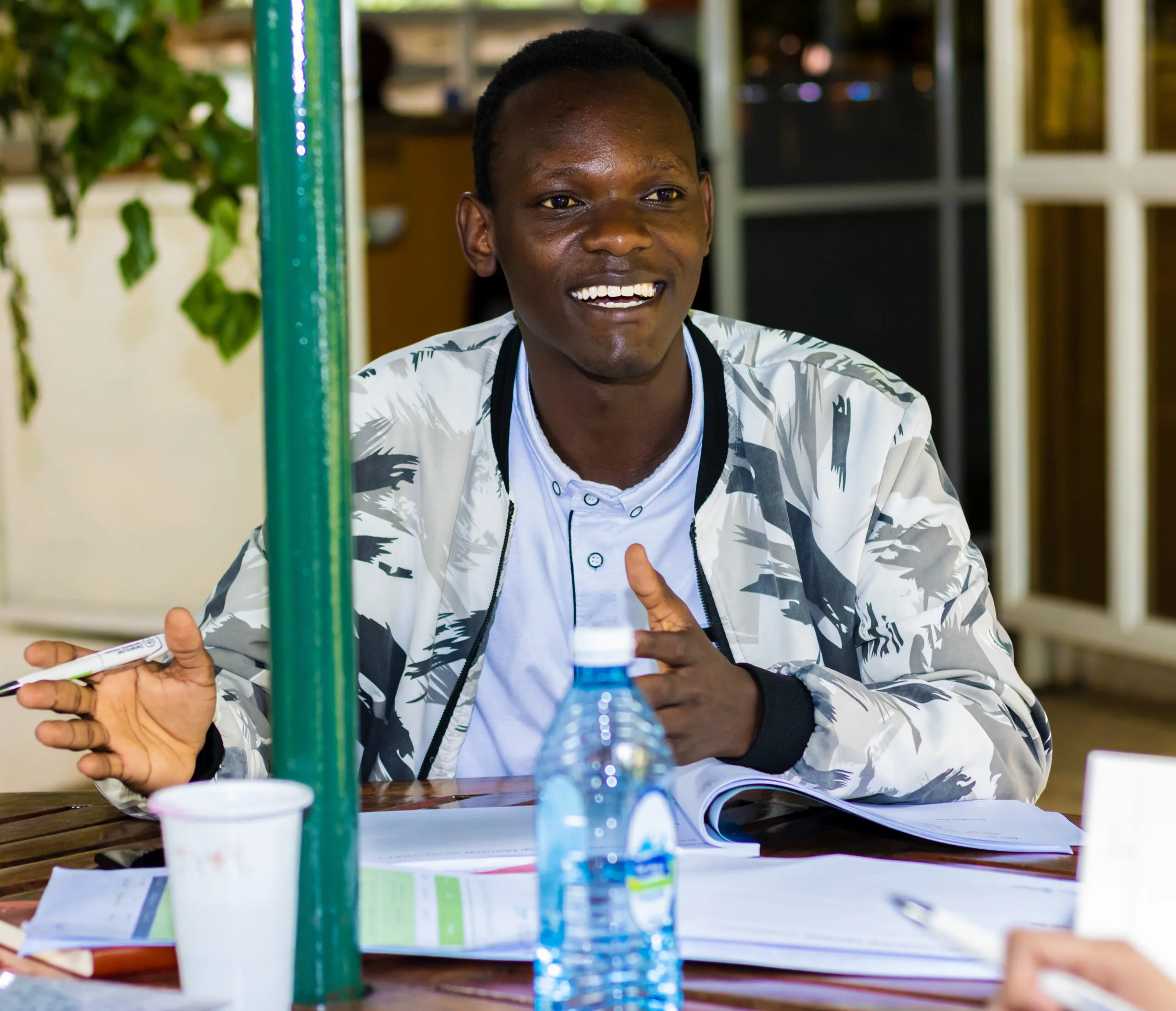 A young entrepreneur smiling and gesturing while talking at a table with documents during an event in Kenya, Thousand Young Entrepreneurs 2024